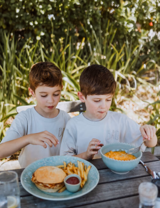 Two young boys in light blue shirts sit at a wooden table outdoors, sharing a meal of burgers, fries, and macaroni in bowls.